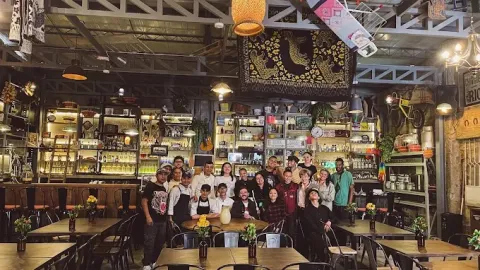 Interior de un bar o restaurante de estilo industrial y ecléctico, con estanterías llenas de botellas, objetos decorativos, instrumentos musicales y plantas. En el centro, un grupo numeroso de personas posa unido y sonriente frente a la barra, algunos abrazándose o inclinándose hacia la cámara. 