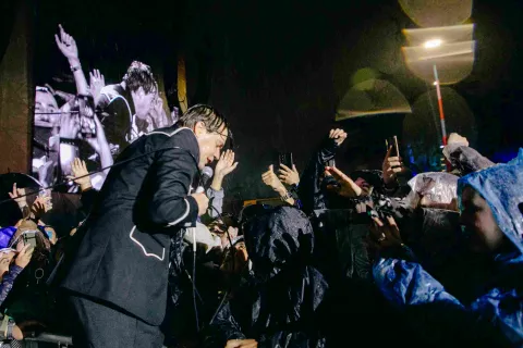 Cantante bajo la lluvia se acerca al público, que levanta las manos y celulares mientras disfrutan el concierto.