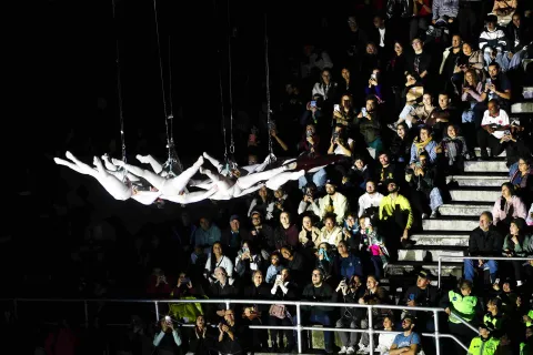 Espectáculo en vivo con acróbatas vestidos de blanco suspendidos horizontalmente en el aire, mientras un público numeroso observa desde gradas, muchos grabando con sus celulares en un ambiente nocturno.