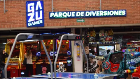 Entrada a un parque de diversiones en un centro comercial con máquinas arcade, mesas de juego y letrero luminoso de “Gazu Games”.