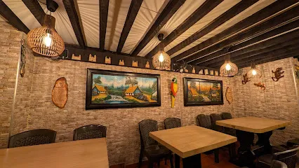  Interior de un restaurante con ambiente cálido y tradicional, decorado con paredes de ladrillo y vigas de madera expuestas en el techo. Varias mesas de madera con sillas tejidas están organizadas de forma ordenada, iluminadas por lámparas colgantes de luz cálida. En las paredes se exhiben cuadros de paisajes y elementos decorativos artesanales.