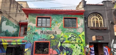  Pasillo interior de un bar o café alternativo con iluminación en tonos azules y morados. Las paredes están cubiertas de carteles, ilustraciones y grafitis, creando un ambiente urbano y artístico. A ambos lados, grupos de personas conversan sentadas en sillas y mesas pequeñas, algunas con bebidas.