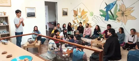 Interior de un espacio acogedor donde un grupo de personas participa en una actividad guiada. A la izquierda, un hombre de pie habla y gesticula frente al grupo. Los asistentes están sentados en el suelo y en cojines alrededor de mesas bajas con teteras, tazas y elementos naturales, sugiriendo una experiencia de infusiones o bienestar.