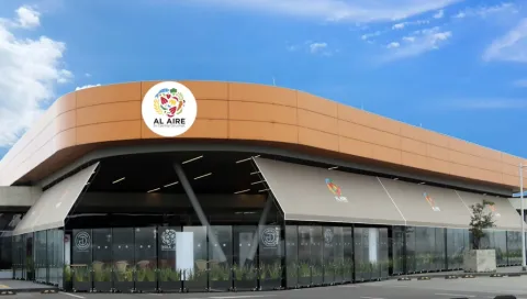  La imagen muestra un moderno espacio comercial con un diseño arquitectónico elegante y contemporáneo, identificado por el letrero “Al Aire” en su parte superior. La estructura se distingue por su forma curva y envolvente, revestida en un tono cálido similar al cobre, que contrasta armoniosamente con el cielo azul y las nubes dispersas del fondo.