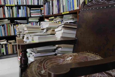 A stack of books behind a wooden chair. In the background is a wooden bookcase overflowing with books.