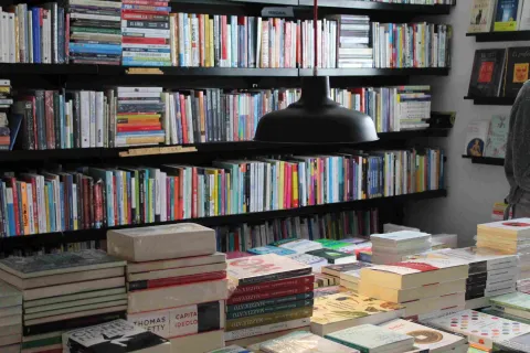 Mesa con pilas de libros bajo una lámpara negra, frente a estanterías llenas de títulos en una librería.
