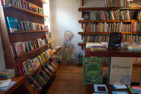 Sección infantil de una librería con estanterías de madera llenas de libros ilustrados, una mesa de exhibición y una figura de El Principito en el fondo.