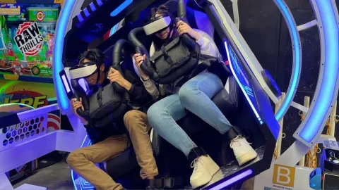 Dos personas usan visores de realidad virtual en una atracción interactiva, sujetas a asientos con arneses dentro de una máquina iluminada.