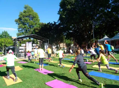Grupo de personas realizando una clase de ejercicio al aire libre sobre tapetes de colores en un parque. 