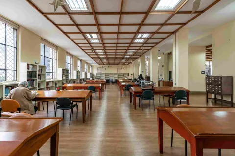 La imagen muestra una sala de lectura amplia y silenciosa, con mesas de madera alineadas y estanterías llenas de libros a lo largo de las paredes. La luz natural entra por grandes ventanas, creando un ambiente tranquilo donde algunas personas leen o estudian.