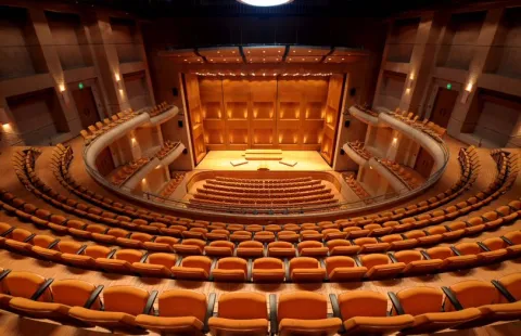 Modern auditorium with an illuminated stage and rows of orange seats arranged in a semicircular layout.