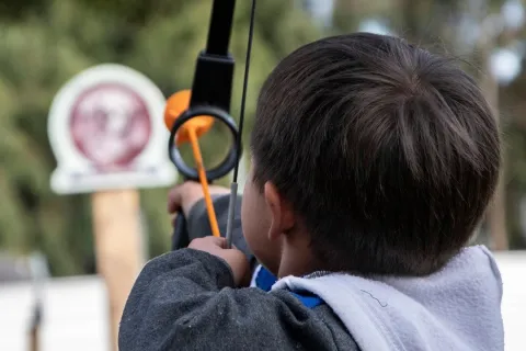 Niño practica tiro con arco apuntando hacia una diana en una actividad al aire libre.