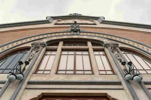 La imagen muestra un detalle de la fachada de un edificio histórico, donde destacan un gran arco decorativo y ventanales enmarcados por elementos ornamentales. Esculturas y lámparas clásicas refuerzan su estilo artístico, mientras el ángulo en contrapicado resalta la altura y elegancia de la construcción. 