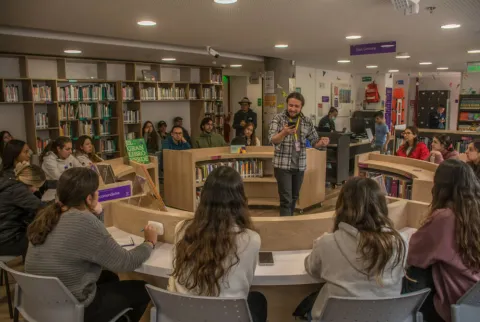 Grupo de personas participa en una actividad en biblioteca, escuchando a un facilitador en el centro del espacio.