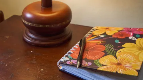  La imagen muestra un cuaderno de espiral con diseño floral colorido, sobre el cual descansa un bolígrafo negro. Está colocado sobre una mesa de madera junto a la base de una lámpara, creando una escena íntima y tranquila que sugiere escritura o reflexión.