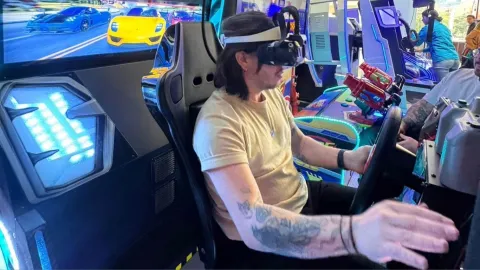 Persona usando visor de realidad virtual mientras maneja un simulador de conducción en una máquina arcade con luces y pantalla de autos.