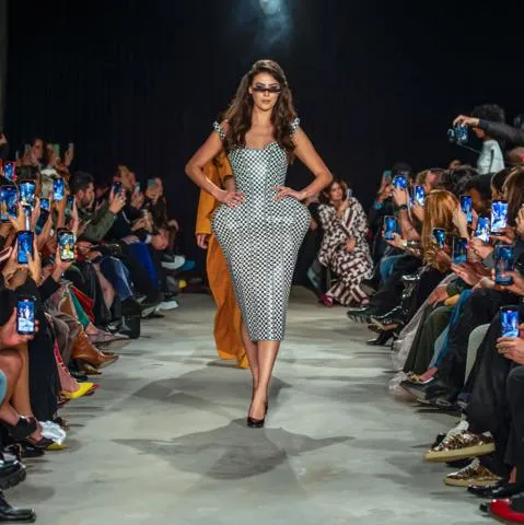  Modelo desfila en pasarela con vestido ajustado a cuadros, mientras el público toma fotos a ambos lados.