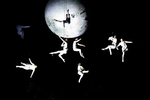  Espectáculo aéreo con varios acróbatas vestidos de blanco suspendidos en el aire alrededor de una gran luna iluminada, creando una escena poética en un fondo oscuro.