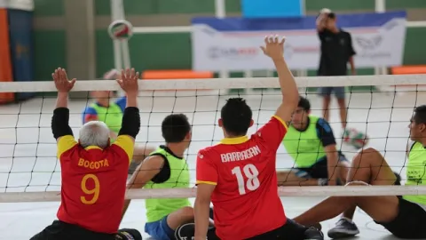 Personas jugando voleibol sentado en una cancha, con participantes levantando las manos frente a la red.