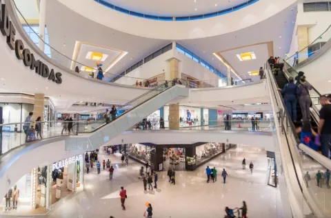 La imagen muestra el interior de un centro comercial de varios niveles, con una arquitectura amplia y luminosa. Escaleras eléctricas conectan los pisos, mientras personas transitan entre tiendas y zonas de comida. La iluminación cálida y los espacios abiertos generan una sensación de dinamismo y modernidad.