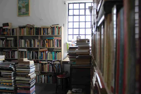 Interior de librería con estanterías llenas y pilas de libros junto a una ventana.