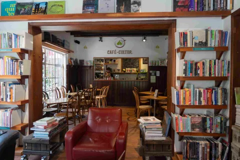 Interior de una librería con café, rodeada de estanterías de libros, mesas de madera y un mostrador al fondo bajo el letrero “Café-Cultor”.