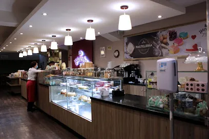 Interior de una pastelería moderna y bien iluminada, con vitrinas de vidrio que exhiben una variedad de postres y productos de repostería. El espacio cuenta con un largo mostrador de madera, lámparas colgantes de luz blanca y paredes decoradas con imágenes de dulces.