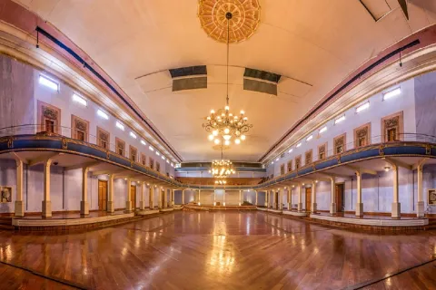 La imagen muestra el interior de un teatro o salón amplio y elegante, con un piso de madera brillante y balcones laterales de estilo clásico. Grandes lámparas colgantes iluminan el espacio, resaltando los detalles arquitectónicos y creando una atmósfera cálida y majestuosa.