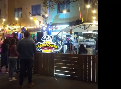 Escena nocturna en una zona gastronómica al aire libre, con varias personas reunidas frente a un puesto de comida llamado “La Berraquera”. El lugar está rodeado por una cerca de madera y decorado con luces colgantes y banderines de colores.