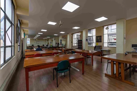 La imagen muestra una sala de lectura amplia y ordenada, con mesas de madera y sillas distribuidas en filas. La luz natural entra por grandes ventanas, complementada por iluminación en el techo.