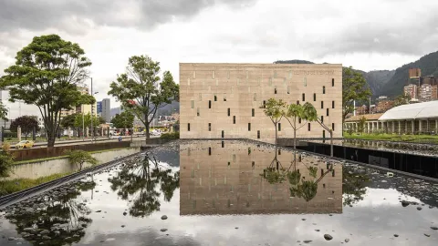 La imagen muestra un edificio moderno de piedra clara reflejado en un espejo de agua en primer plano, creando una composición simétrica y serena. Alrededor, árboles y elementos urbanos acompañan la escena, mientras el cielo nublado y las montañas al fondo refuerzan una atmósfera tranquila y contemplativa.