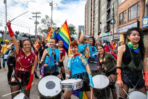  Grupo de personas marchando en un desfile del orgullo LGBTI+ por una calle urbana, tocando tambores y sonriendo; llevan vestuarios coloridos, maquillaje llamativo y banderas arcoíris, mientras una multitud los acompaña a ambos lados de la vía.