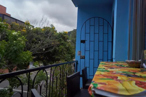 La imagen muestra un balcón acogedor con paredes azules y una mesa cubierta con un mantel colorido. Una baranda de hierro enmarca la vista hacia árboles y una calle tranquila, bajo un cielo nublado. El espacio transmite calma y un ambiente íntimo al aire libre.