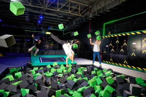 Personas saltan y juegan en una piscina de cubos de espuma, mientras una de ellas realiza una voltereta en un parque de trampolines interior.