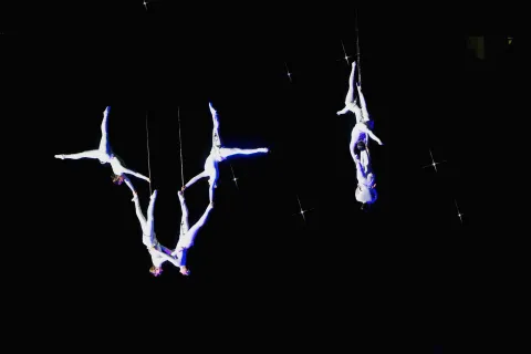 Acróbatas vestidos de blanco realizan figuras suspendidos en el aire con arneses, formando composiciones simétricas sobre un fondo negro con destellos de luz que evocan estrellas.