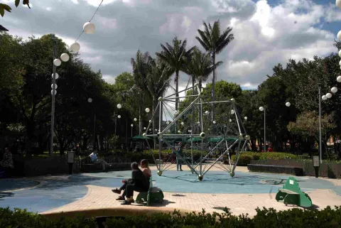 Área de juegos con una estructura metálica para escalar, rodeada de árboles y personas sentadas en bancas. 
