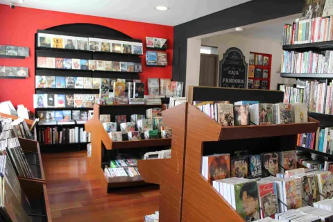  Interior de librería con estanterías de madera llenas de libros y cómics, con pared roja y exhibidores organizados.
