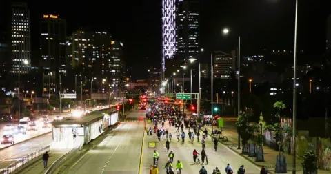 Ciclistas recorren una vía principal de Bogotá durante la noche, con edificios iluminados y tráfico alrededor.