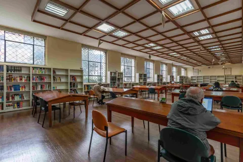 La imagen muestra una sala de lectura amplia y bien iluminada, con mesas de madera y estanterías llenas de libros a lo largo de las paredes. Varias personas leen o trabajan en silencio, creando un ambiente tranquilo y concentrado. La luz natural que entra por las ventanas y el techo geométrico aportan calidez y orden al espacio.