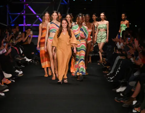 Grupo de modelos desfila en pasarela con vestidos coloridos y estampados, ante público a ambos lados.