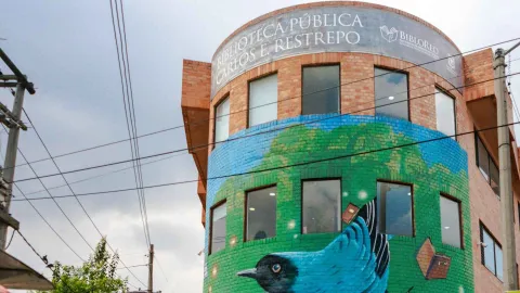 Facade of the Carlos E. Restrepo Public Library with a colorful mural of a bird and books, standing out against the urban backdrop under a cloudy sky.