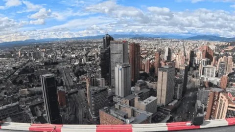  Vista panorámica de una ciudad con numerosos edificios altos y avenidas amplias, observada desde gran altura. El horizonte muestra montañas lejanas bajo un cielo parcialmente nublado.