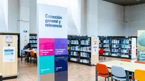 Interior de una biblioteca pública con estanterías llenas de libros y mesas de estudio. En primer plano, un letrero indica “Colección general y referencia” con áreas como ciencias sociales, ciencias aplicadas, bellas artes y literatura.