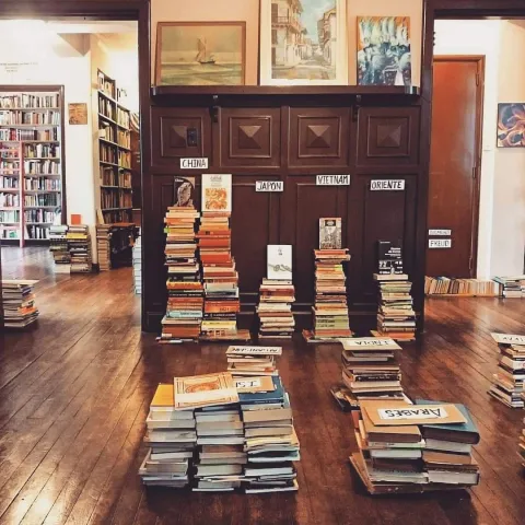 Interior of the bookstore with stacks of books organized by subject on a wooden floor and shelves in the background.