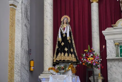 Escultura de la Virgen vestida de negro con detalles dorados, ubicada entre columnas y un fondo de cortinas rojas, acompañada de flores y velas.