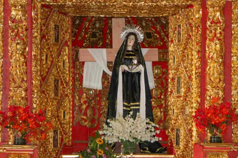 Altar religioso decorado en dorado y rojo, con una escultura de la Virgen de los Dolores frente a una cruz, rodeada de flores y ornamentos barrocos.