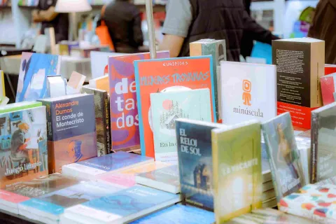 An exhibition table showcasing a diverse selection of books and novels at a literary fair, surrounded by visitors and independent publishers in a vibrant, creative atmosphere.