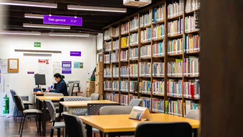  Sala de biblioteca con estanterías llenas de libros y mesas de estudio, donde una persona trabaja en un ambiente tranquilo bajo el letrero “Sala general”.