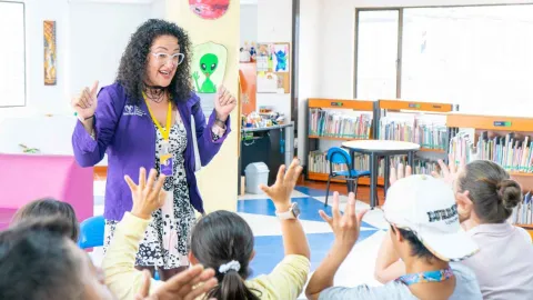 A library facilitator interacts with a group of children raising their hands in a colorful space that encourages reading and learning.