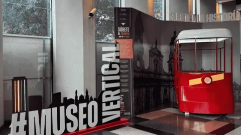  Espacio interior de museo con una exhibición interactiva: un vagón rojo tipo teleférico junto a paneles informativos. En primer plano se lee “#MUSEO VERTICAL”, con elementos gráficos que evocan la historia de la ciudad.